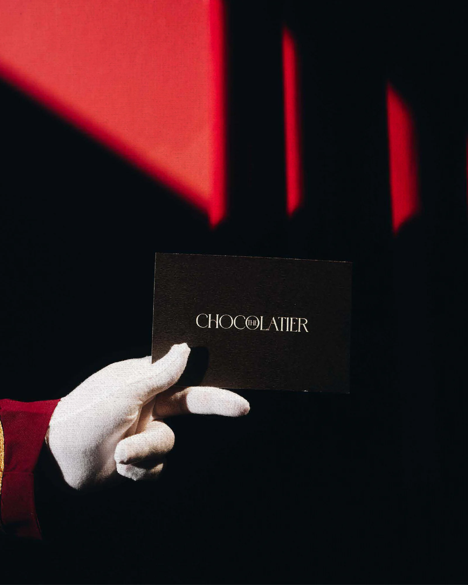 White-gloved hand holding a dark Chocolatier card beneath red light